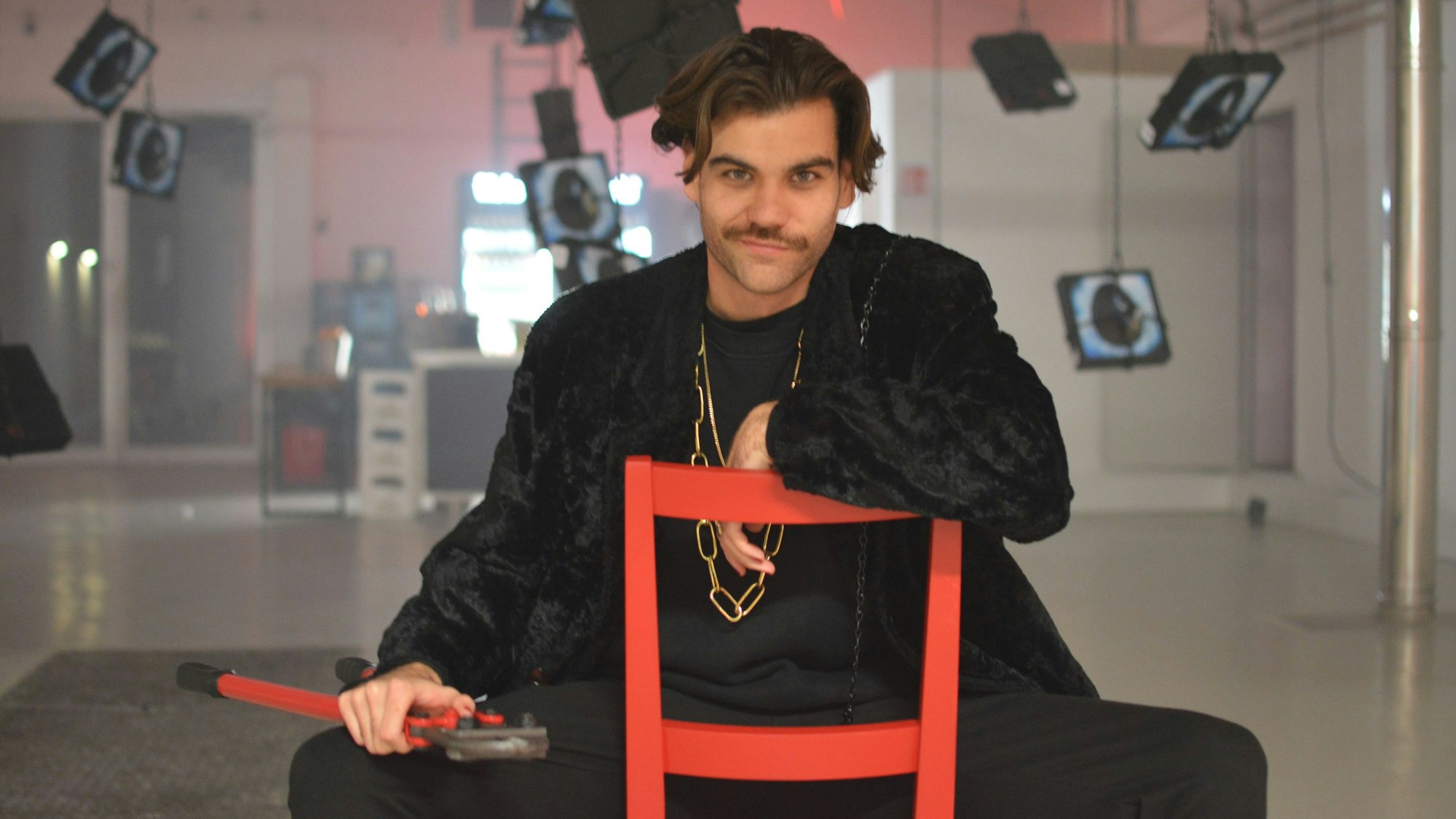 Max Leinfelder sitzt auf einem roten Stuhl, er trägt schwarze Kleidung und hat einen Bolzenschneider in der Hand.
