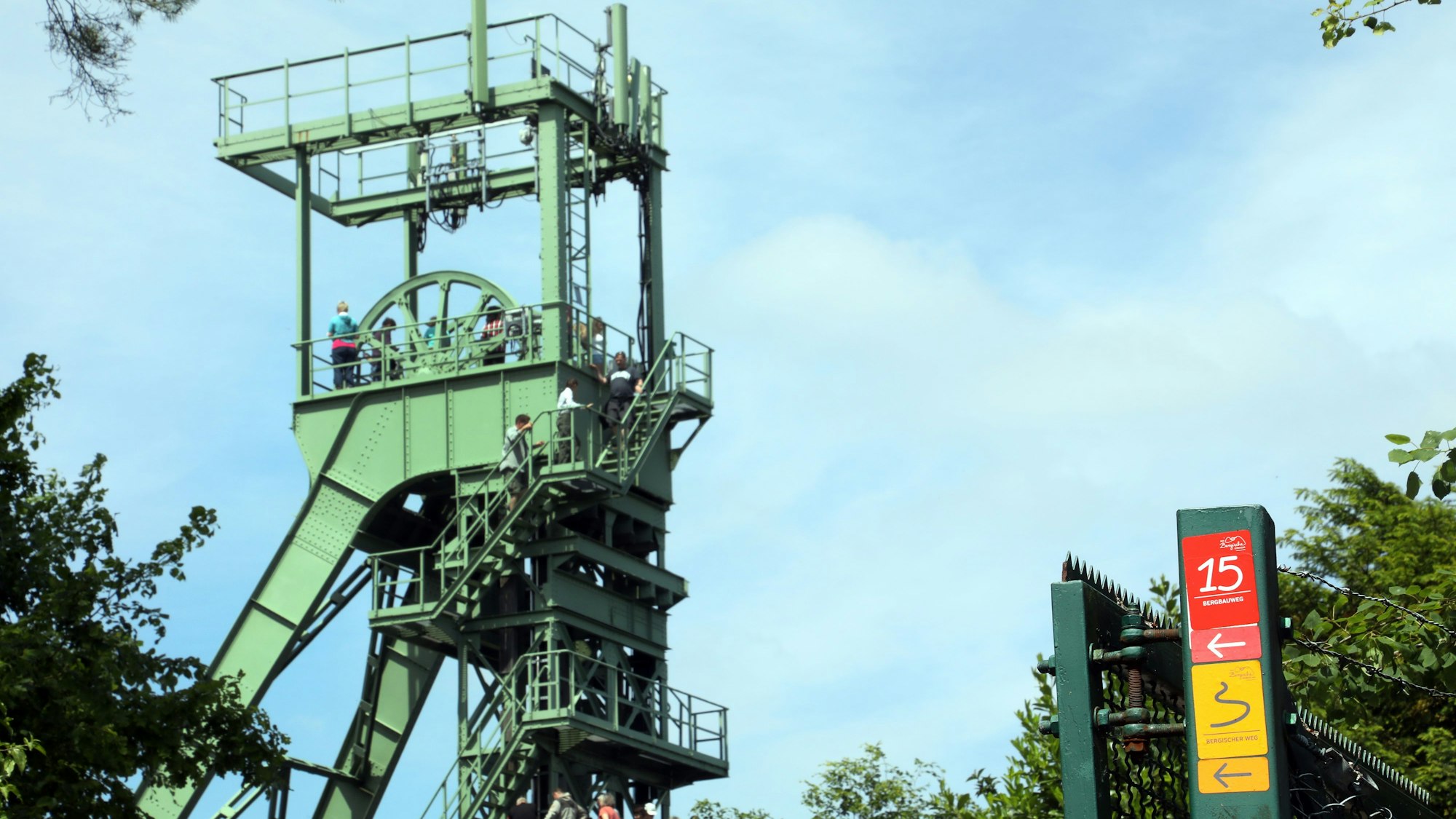 Ein Förderturm ist vor blauem Himmel zu sehen, im Vordergrund eine Wanderwegmarkierung.