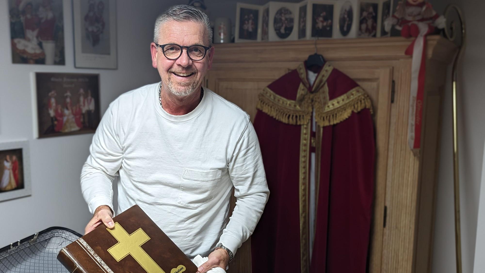 Ein Mann mit weißem Sweatshirt packt eine große Klasse in einen Koffer. Im Hintergrund hängt der Nikolaus-Mantel an einem Schrank.