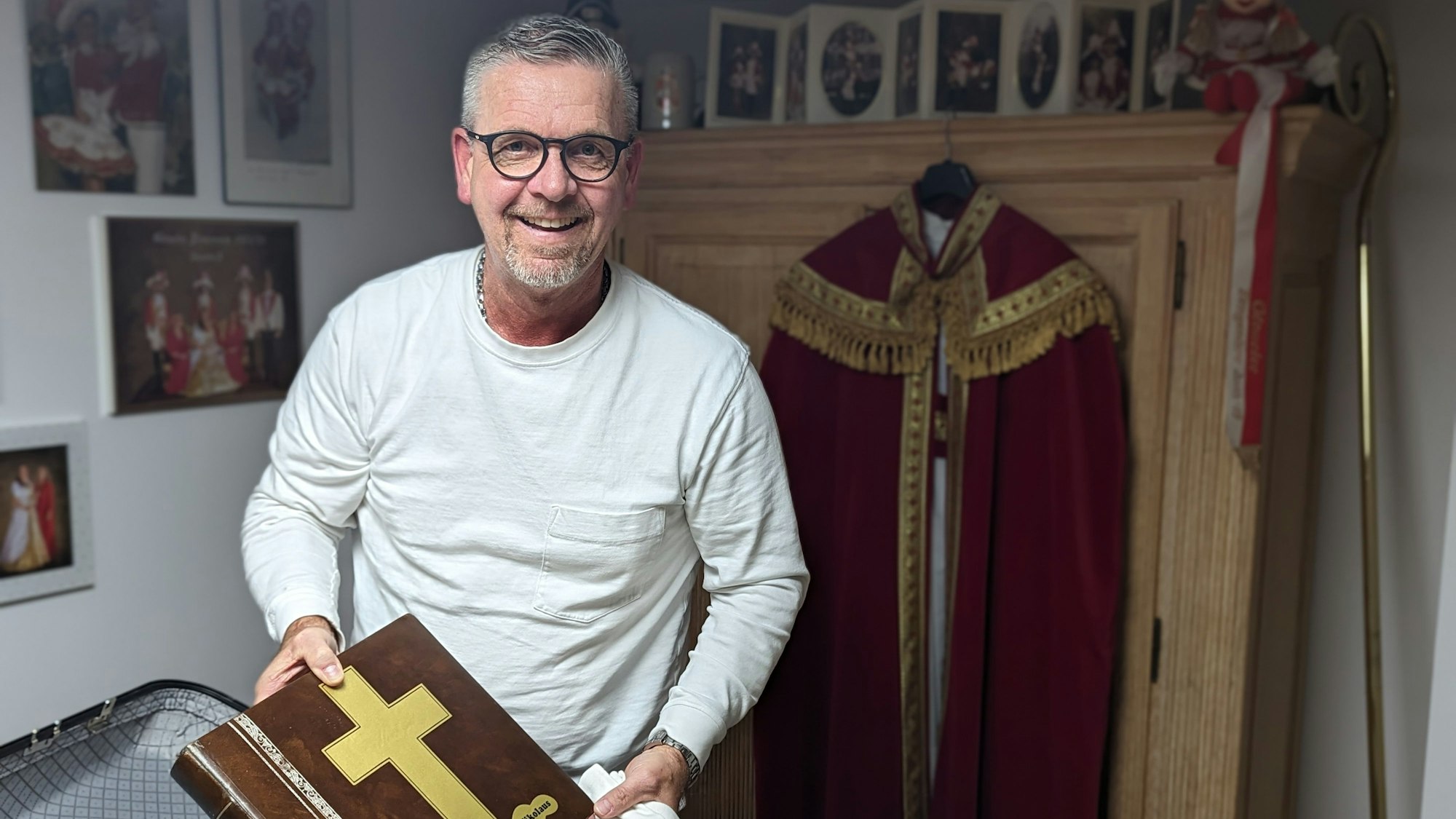 Ein Mann mit weißem Sweatshirt packt eine große Klasse in einen Koffer. Im Hintergrund hängt der Nikolaus-Mantel an einem Schrank.