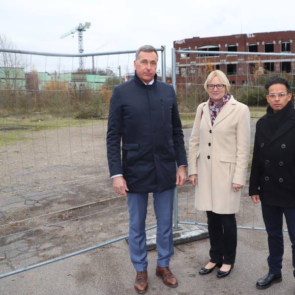 Auf dem Foto sind Wesselings Bürgermeister Ralph Manzke, KVB-Vorstandsvorsitzende Stefanie Haaks und Projektleiter Manuel Franke (v.l.) vor dem Bauzaun der Industriebrache der ehemaligen Norton-Fabrik in Wesseling am Kronenweg zu sehen.