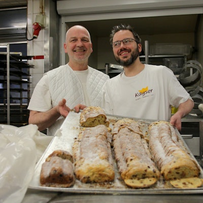 Zwei Bäcker halten ein riesiges Backblech in ihren Händen, darauf liegen vier Christstollen.