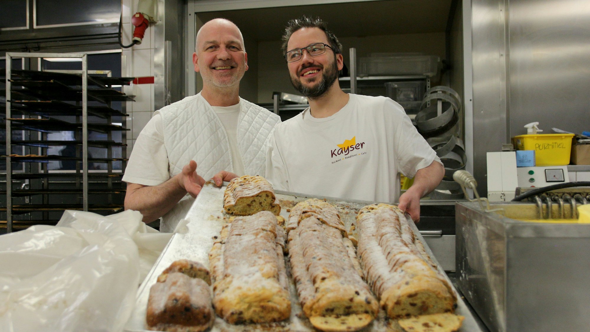 Zwei Bäcker halten ein riesiges Backblech in ihren Händen, darauf liegen vier Christstollen.