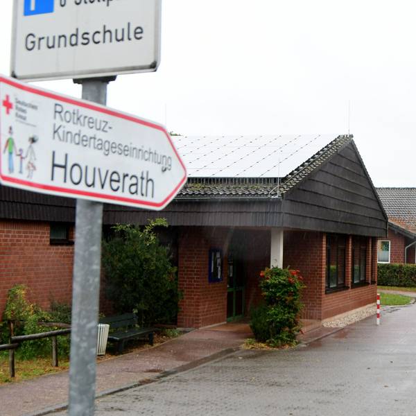 Auf einem Wegweiser steht "Rotkreuz-Kindertageseinrichtung Houverath", dahinter ist der Kindergarten zu sehen.