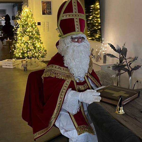 Ein Mann im Nikolauskostüm mit Mantel, Mitra und Rauschebart vor einem hell erleuchteten Weihnachtsbaum.