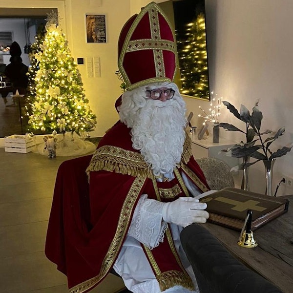 Ein Mann im Nikolauskostüm mit Mantel, Mitra und Rauschebart vor einem hell erleuchteten Weihnachtsbaum.