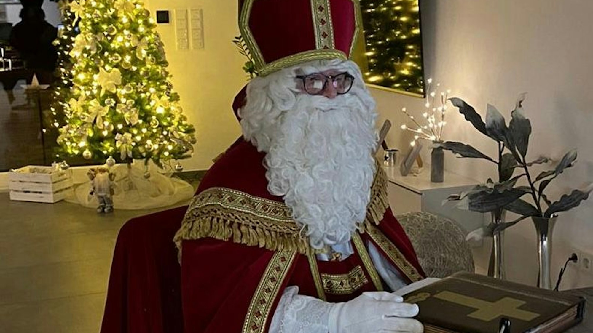 Ein Mann im Nikolauskostüm mit Mantel, Mitra und Rauschebart vor einem hell erleuchteten Weihnachtsbaum.