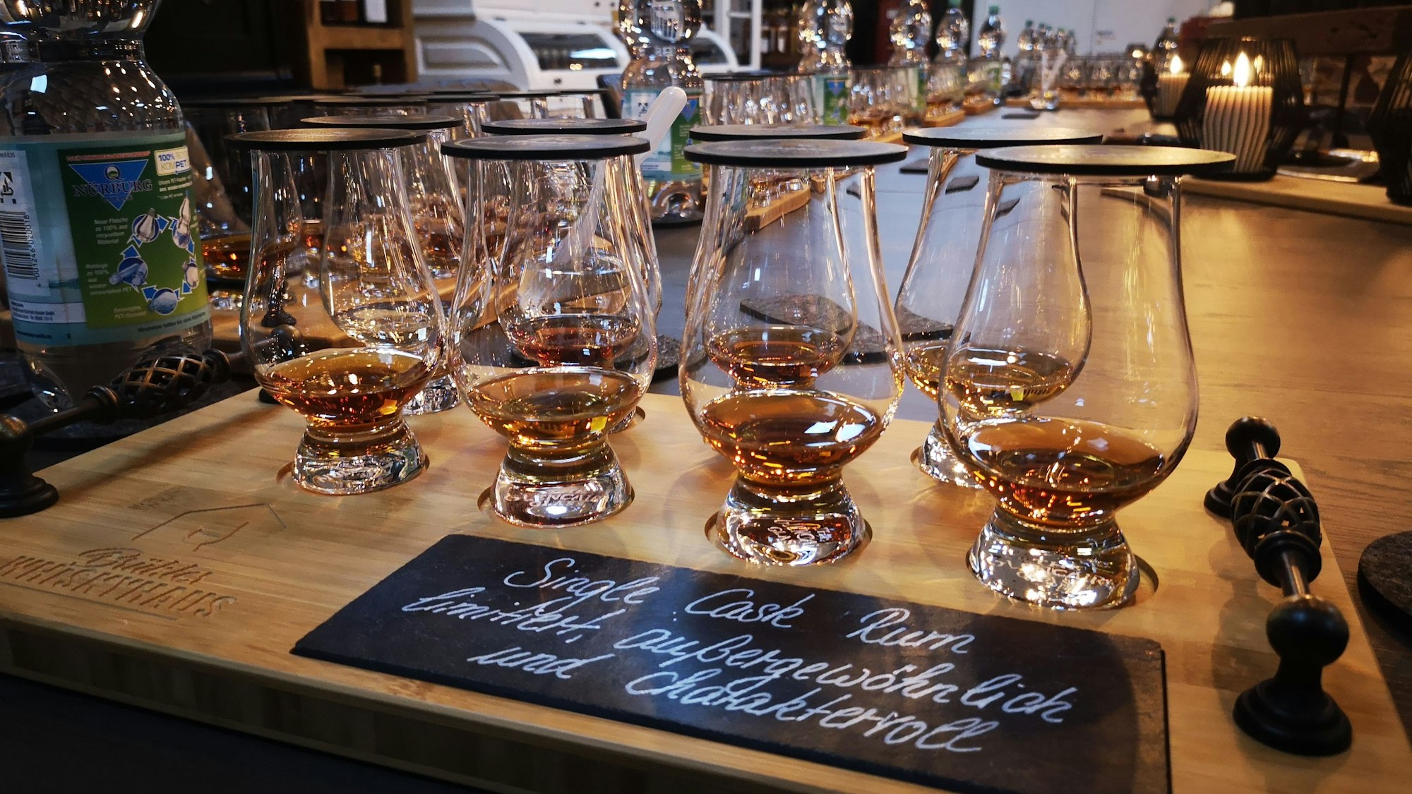 Acht mit Rum gefüllte Gläser stehen auf einem Holzbrett. Deckel verhindern, dass das Aroma der Spirituose entweicht. Auf einer kleinen eingelassenen Kreidetafel steht das Motto des Abens unter „Single Cask Rum“.
