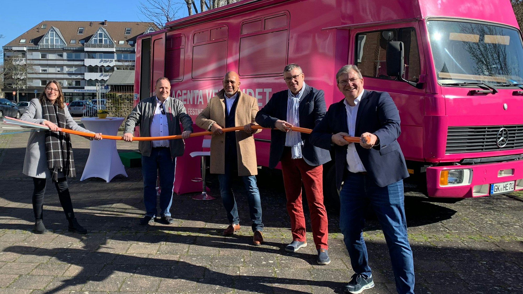 Vertreter der Stadt Niederkassel und der Telekom halten ein dickes Glasfaserkabel in der Hand. Im Hintergrund steht ein magentafarbenes Infomobil der Telekom.