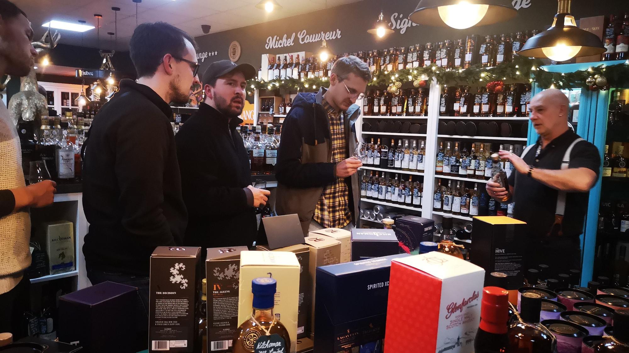 Drei Männer stehen vor einem Tisch und einem Regal mit vielen Whisky-Flaschen. Ein Mitarbeiter hält eine Flasche in der Hand und erzählt von der Sorte, die er gerade zur Verkostung anbietet.