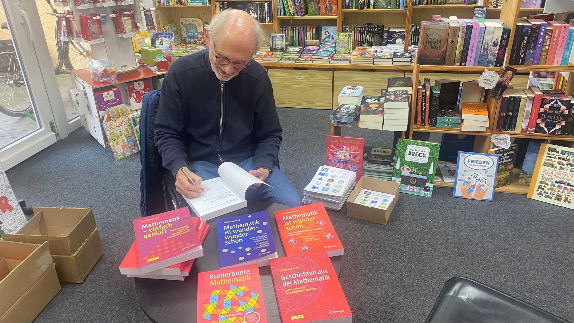 Ein Mann sitzt in einer Buchhandlung am Tisch und schreibt eine Widmung in ein Buch.