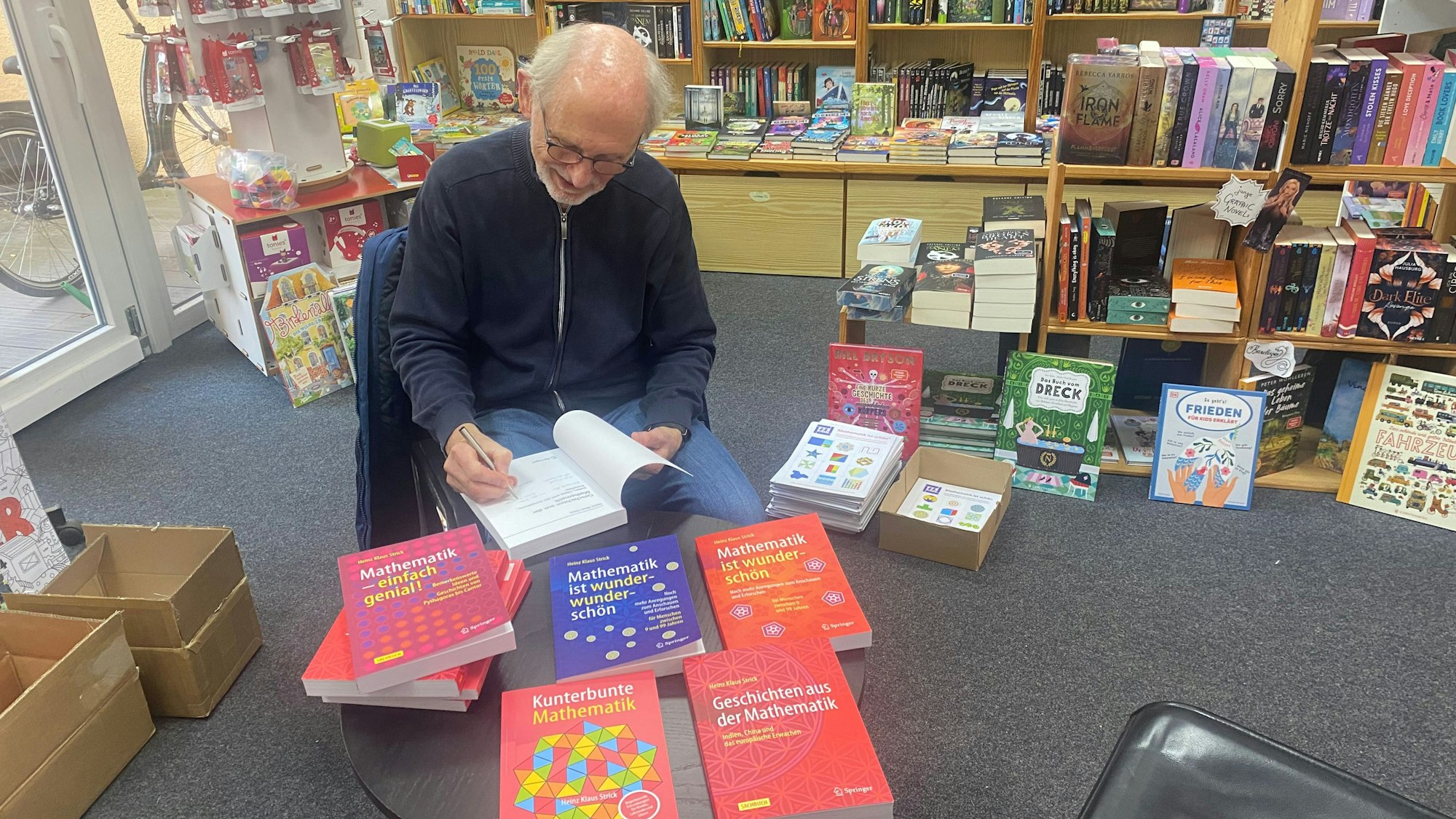 Ein Mann sitzt in einer Buchhandlung am Tisch und schreibt eine Widmung in ein Buch.