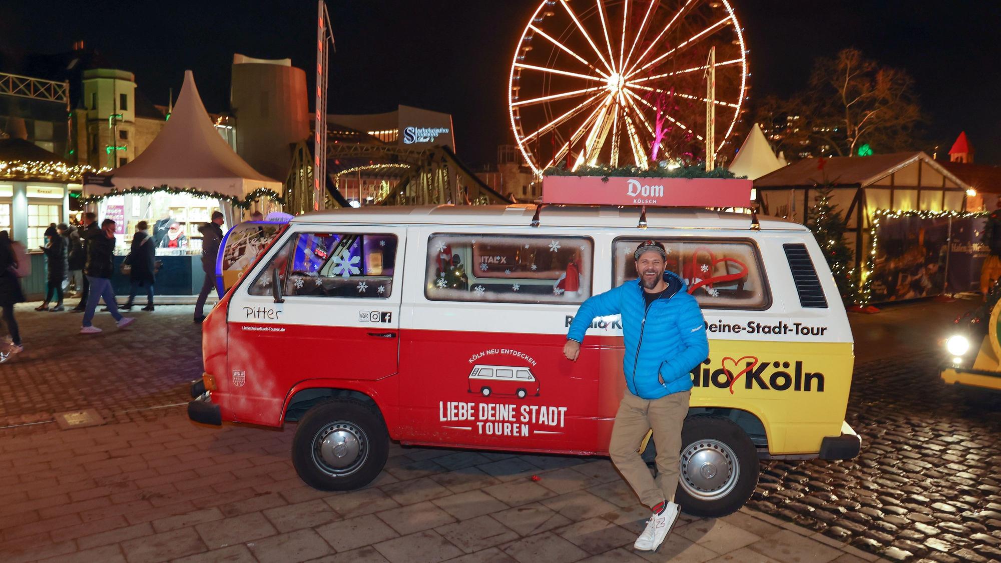 Roman Bendt steht vor dem roten VW-Bulli, hinter ihm das Riesenrad und die Hütten des Hafen-Weihnachtsmarkts.