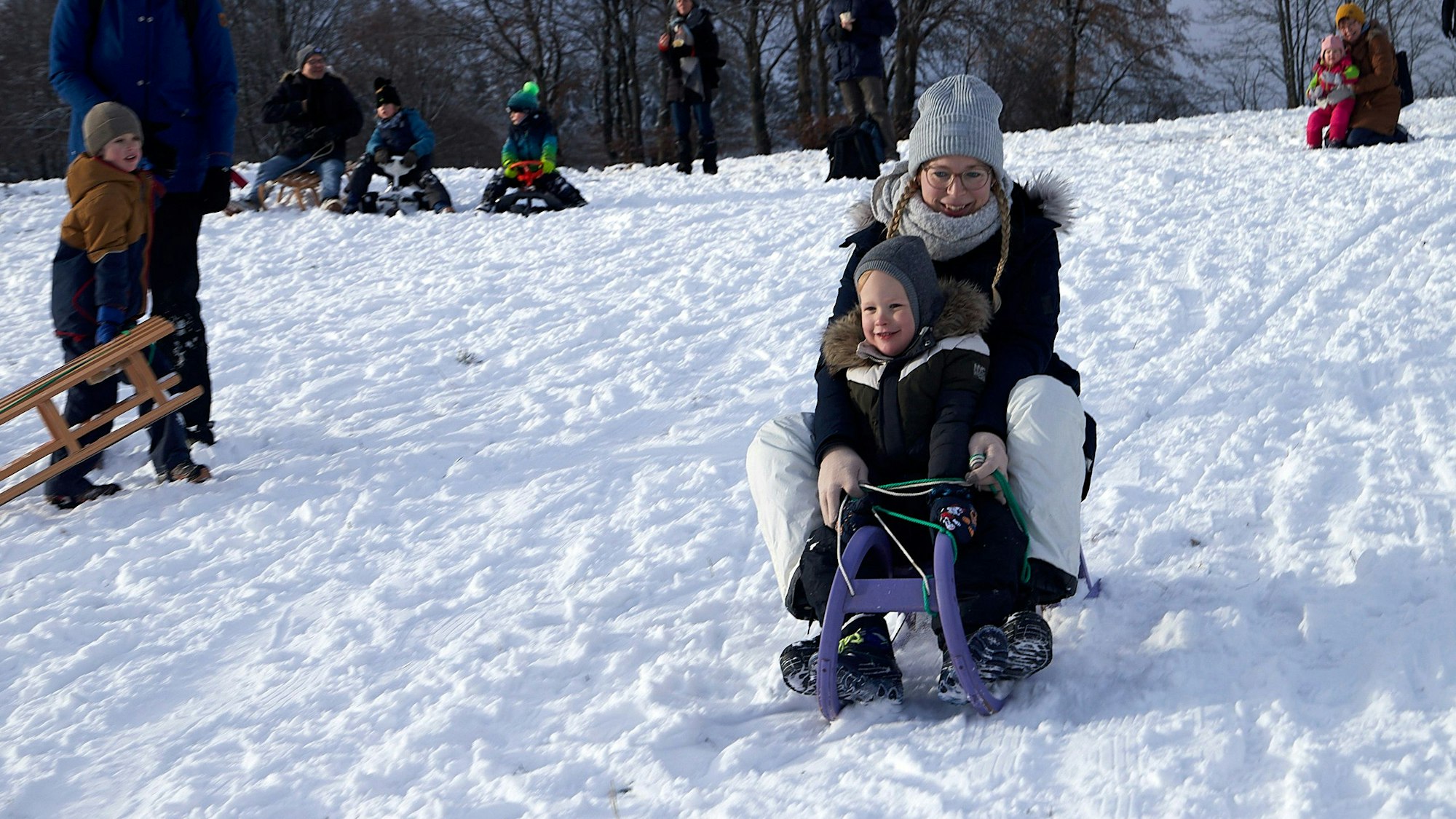 Mutter und Kind rodeln auf einem Kunststoffschlitten den Abhang hinunter.