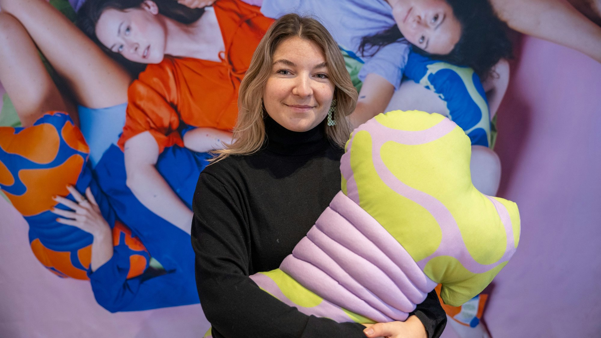 Sanja Zündorf steht mit einem bunten Kissen im Arm vor dem Bild von mehreren Frauen, die inmitten von Kissen liegen.