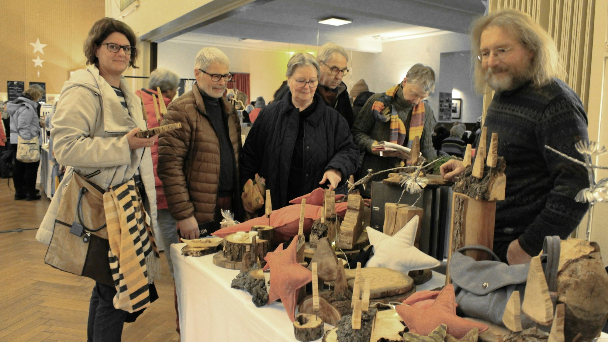 Theo Weidenbach steht hinter einem Tisch, auf dem zahlreiche Sterne, Kerzenhalter oder dargestellte Weihnachtsbäume aus Holz stehen. Besucher schauen sich die Ware an.
