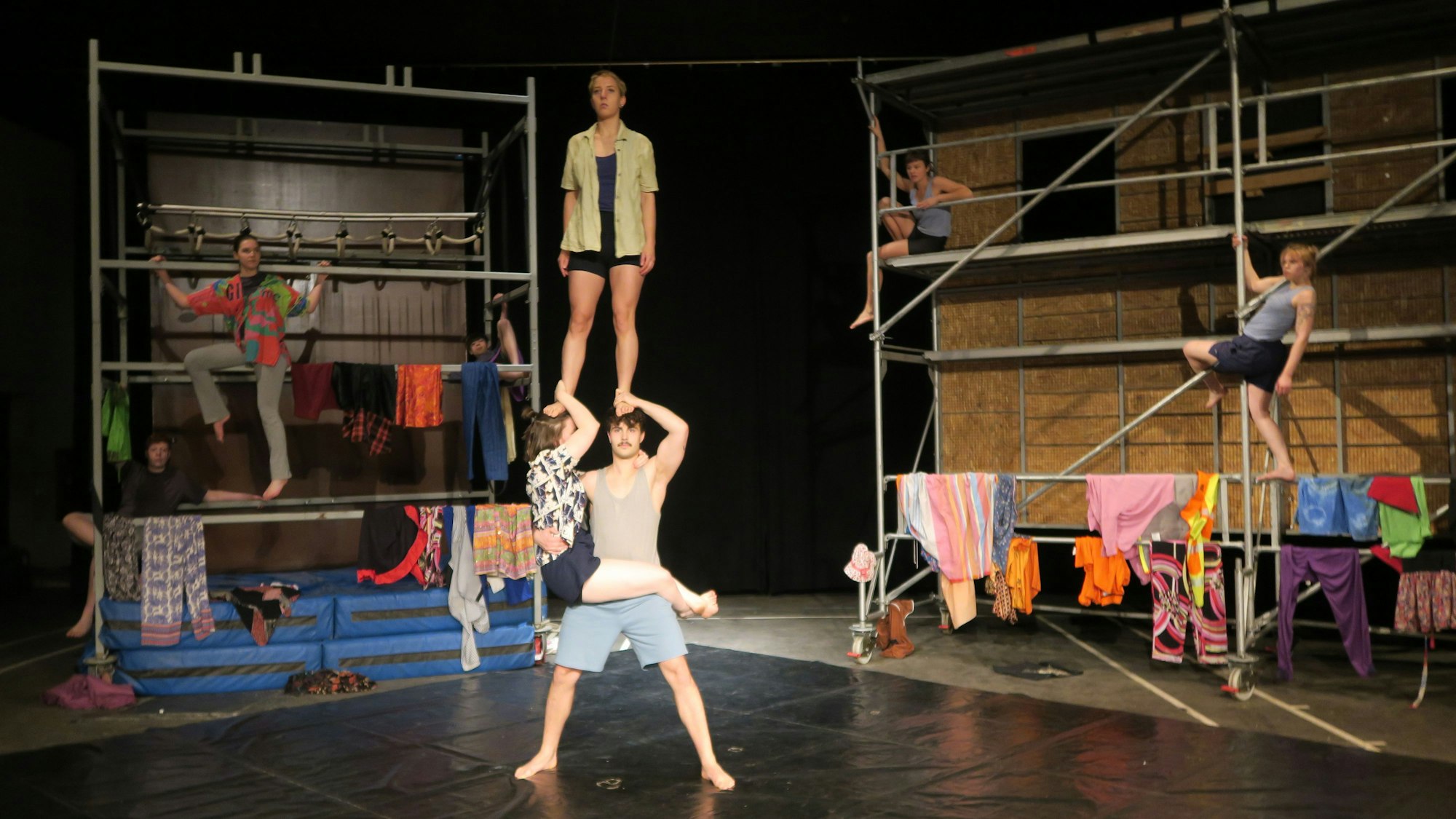 Schauspieler stehen auf einer Bühne, drei bilden eine Art Menschenpyramide, drei weitere posieren auf zwei Baugerüsten.