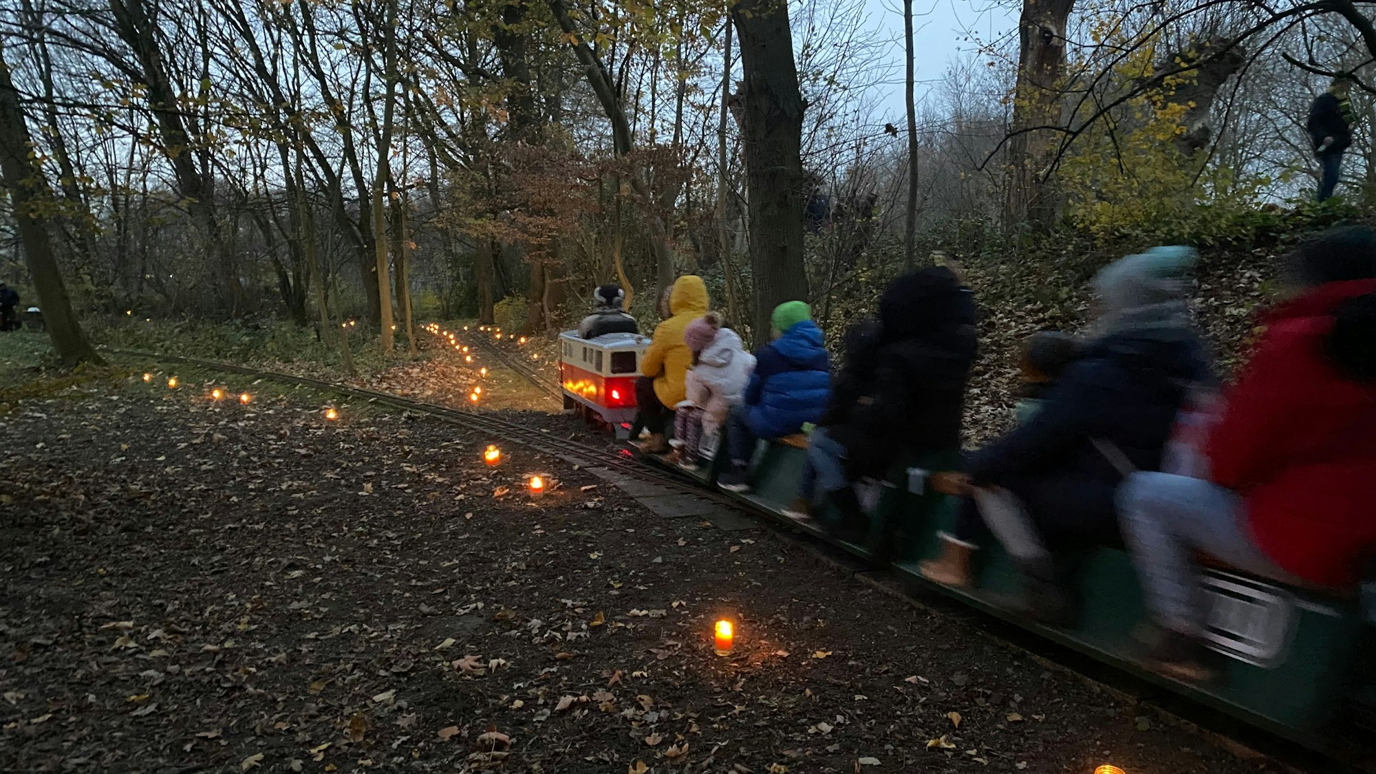 Eine kleine Dampfeisenbahn fährt durch ein Waldstück, darauf sitzen Erwachsene und Kinder. Neben den Gleisen brennen Kerzen.