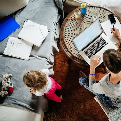Eine Mutter arbeitet an einem Laptop, ihr Kleinkind spielt daneben.