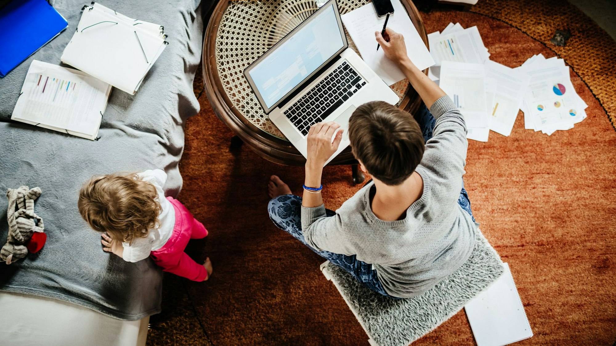 Eine Mutter arbeitet an einem Laptop, ihr Kleinkind spielt daneben.