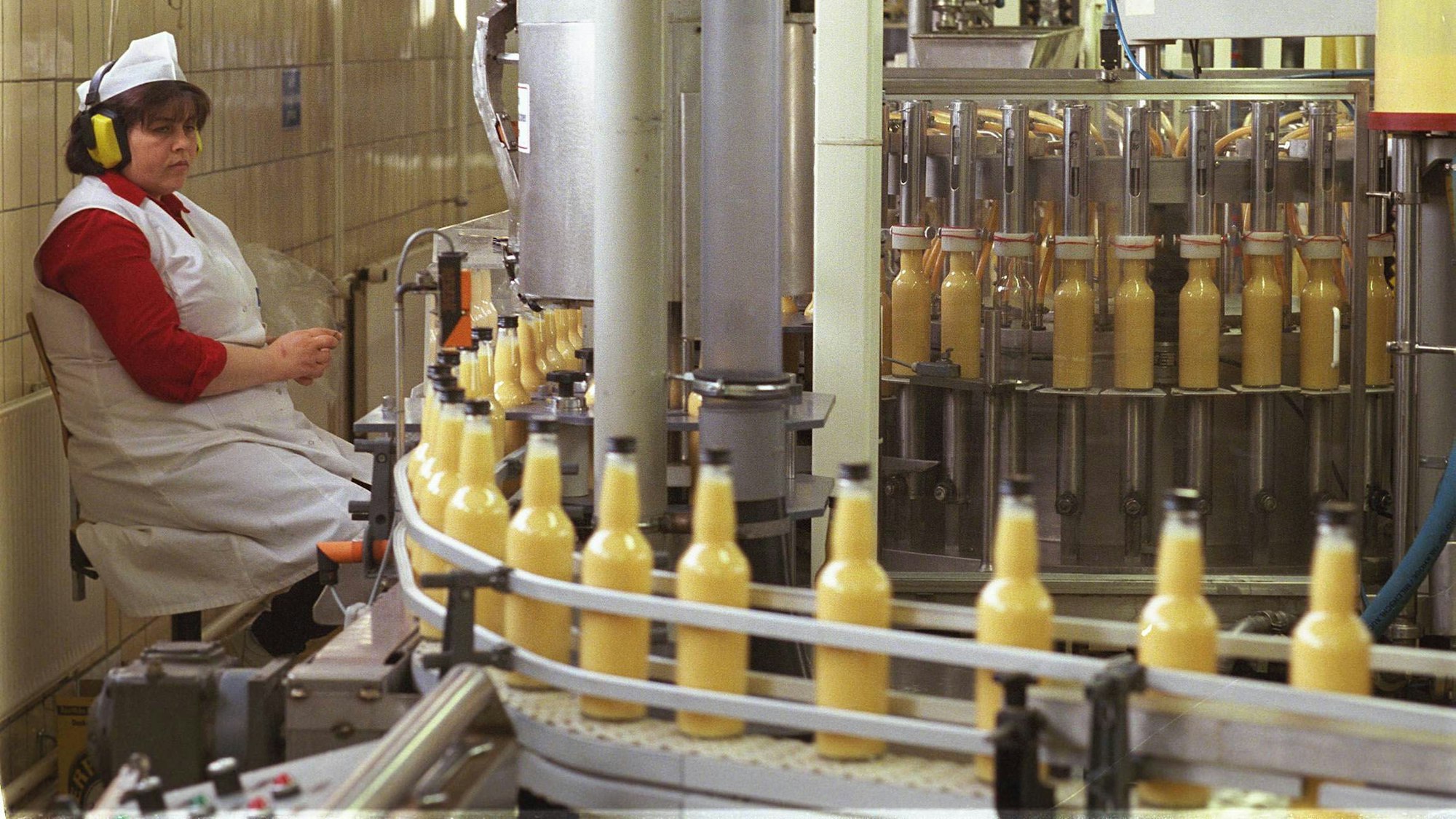 Eine Frau sitzt auf einem Stuhl in der Produktion der Fabrik von Verpoorten in Bonn. Viele Flaschen, befüllt mit Eierlikör, fahren über das Band an ihr vorbei.