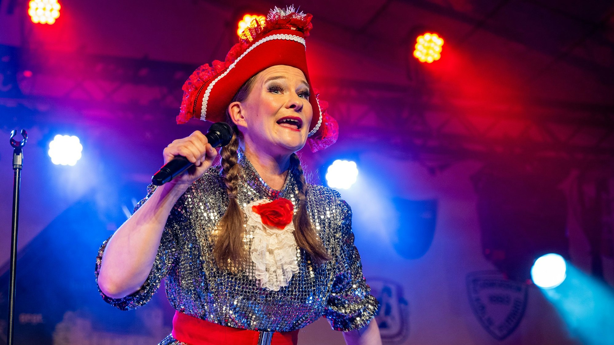 Vera Passy steht in ihrem Kostüm mit silbernem Kleid und roten Hut auf einer Bühne, sie hält ein Mikrofon in der Hand.