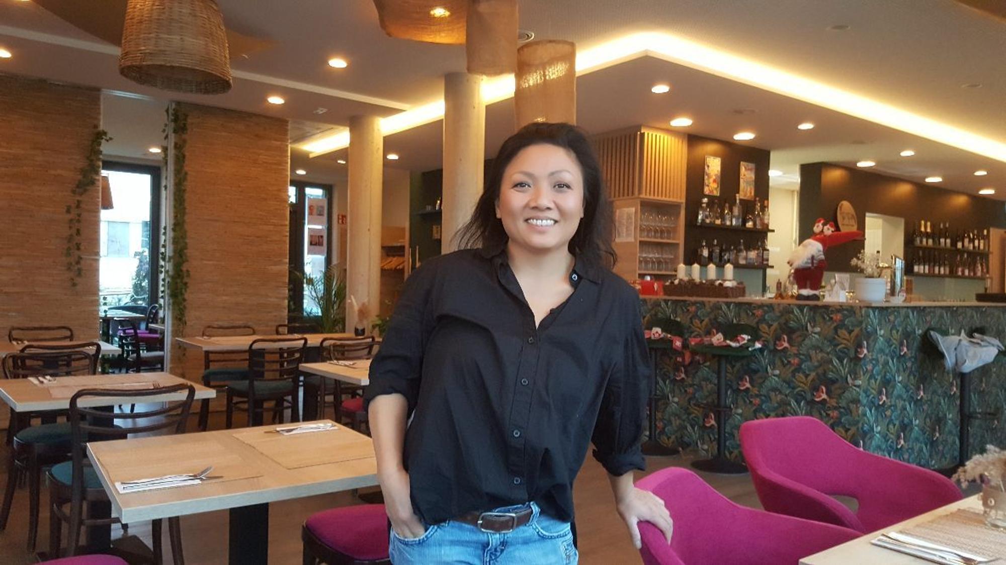 Eine asiatisch aussehende Frau stützt die Hand auf einen Stuhl. Sie steht in einem Restaurant mit vielen hellen Tischen und rosafarbenen Stühlen.