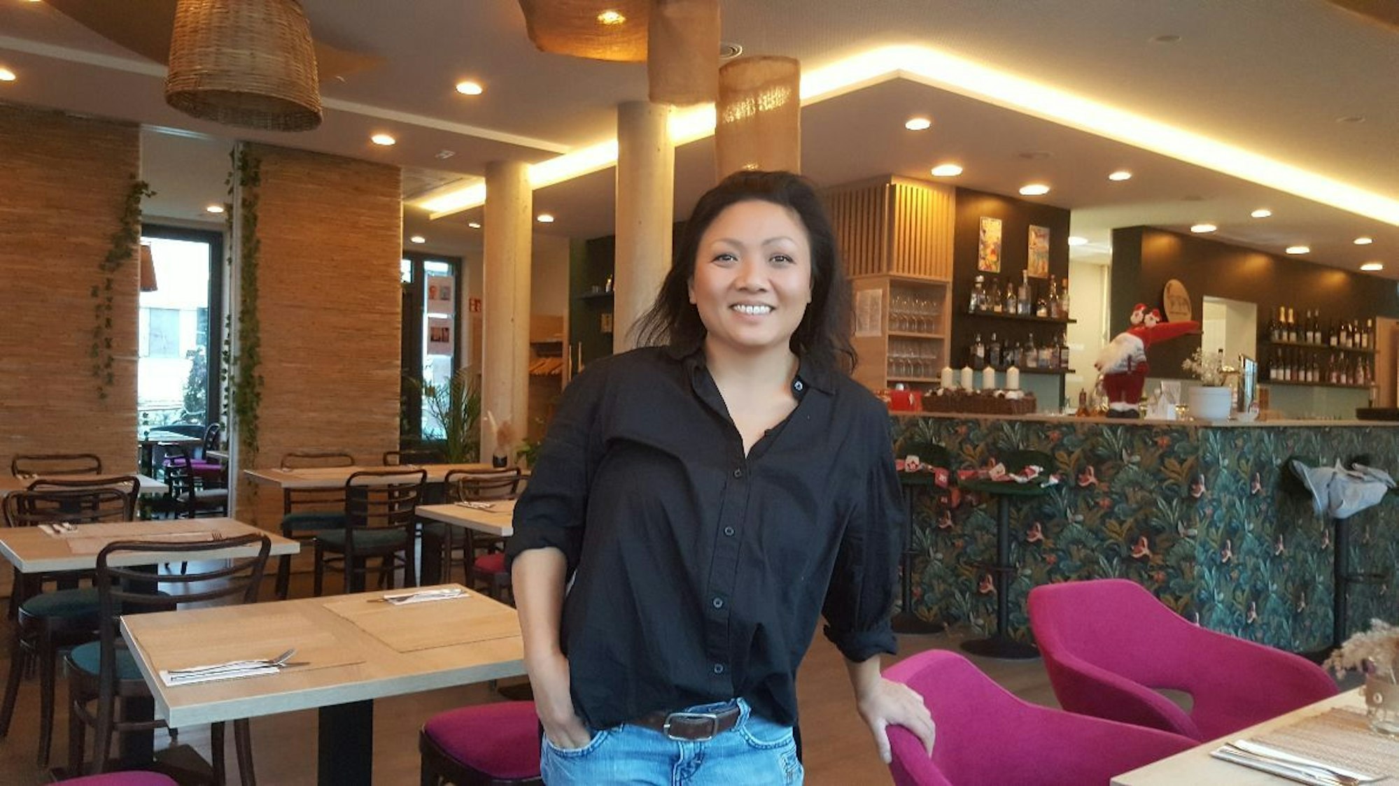 Eine asiatisch aussehende Frau stützt die Hand auf einen Stuhl. Sie steht in einem Restaurant mit vielen hellen Tischen und rosafarbenen Stühlen.