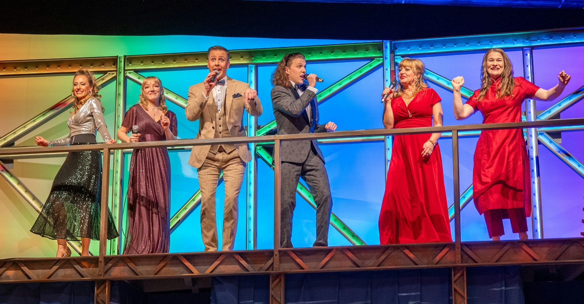 Sechs Sänger stehen auf einer Bühne, rechts tanzt Vera Passy in rotem Kleid.