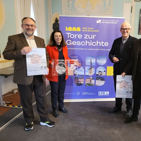 Drei Männer und eine Frau stehen vor einem Plakat mit der Aufschrift 1000 Jahre Abtei Brauweiler - Tore zur Geschichte.