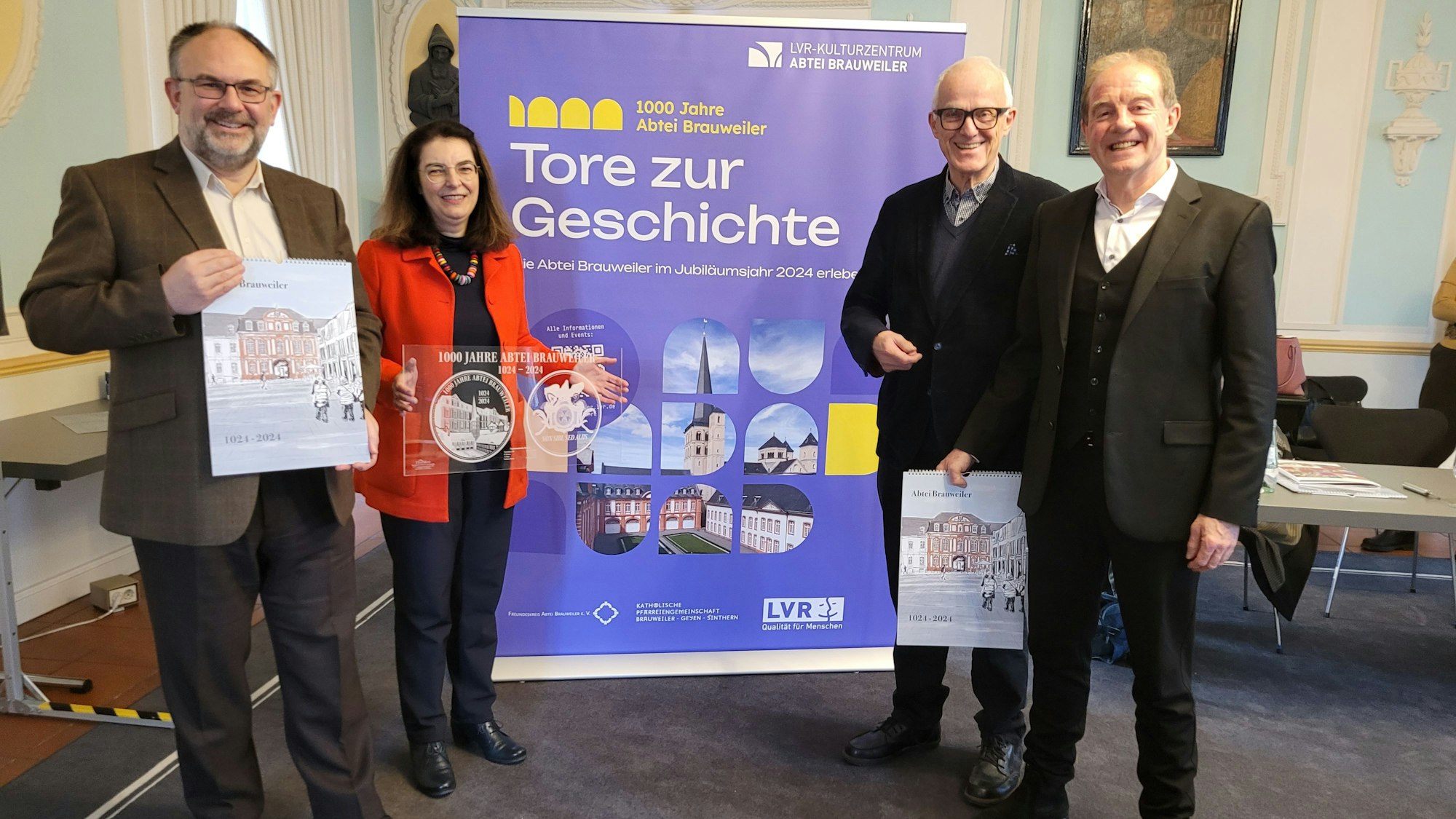 Drei Männer und eine Frau stehen vor einem Plakat mit der Aufschrift 1000 Jahre Abtei Brauweiler - Tore zur Geschichte.
