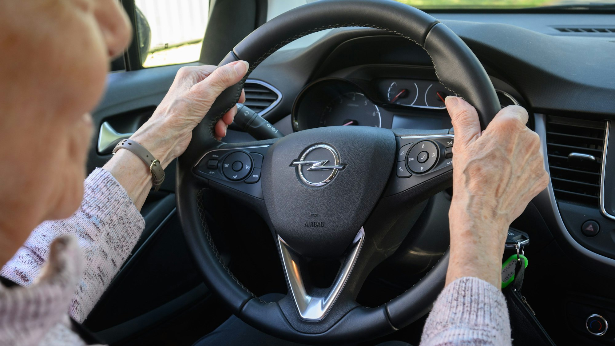 Das Symbolbild zeigt eine Seniorin, die am Steuer eines Autos sitzt. Sie umfasst mit beiden Händen das Lenkrad.