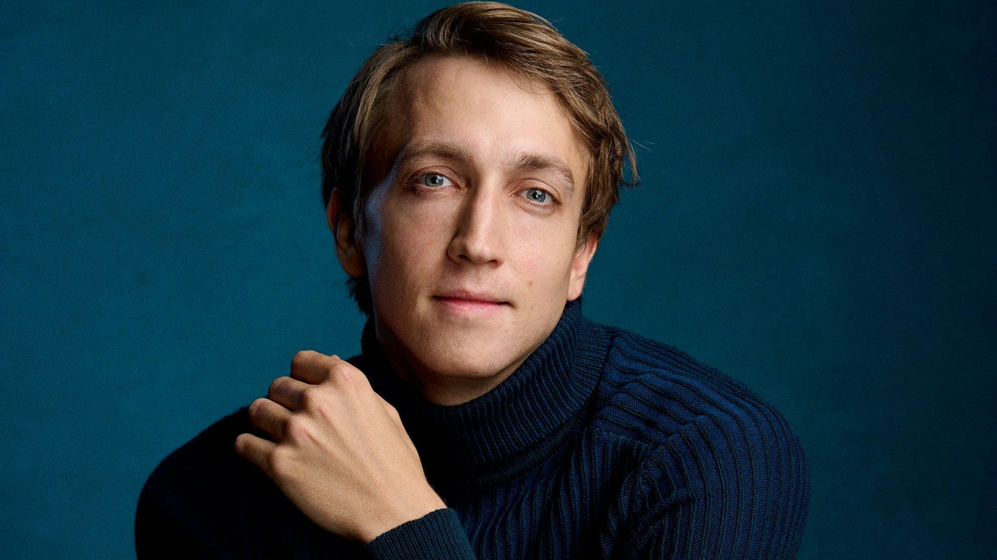 Fabian Müller hat glatte, braune Haare. Er trägt einen dunkelblauen Kragenpullover. Der Hintergrund ist blau.