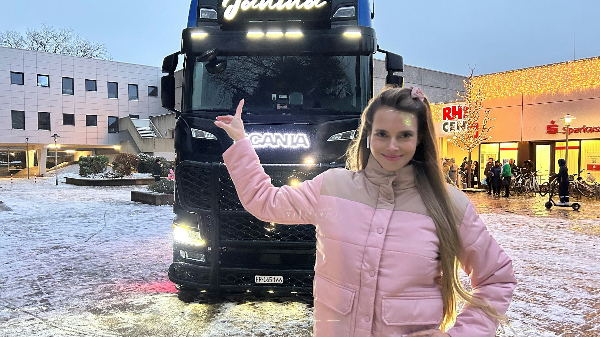 Eine Frau in rosa Jacke zeigt stolz auf einen beleuchteten LKW.