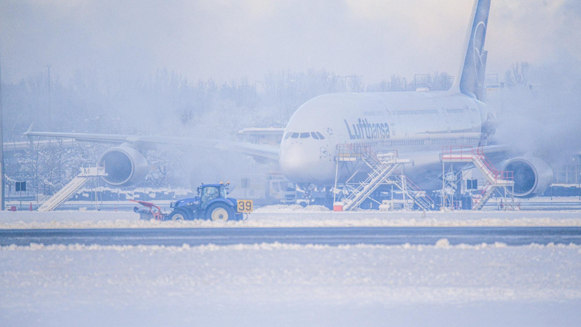 Ein Schneeräumer fährt im Schneetreiben auf dem Flughafen vor einer Maschine entlang. Wegen Eisregen konnten Flugzeuge in München stundenlang nicht starten oder landen.