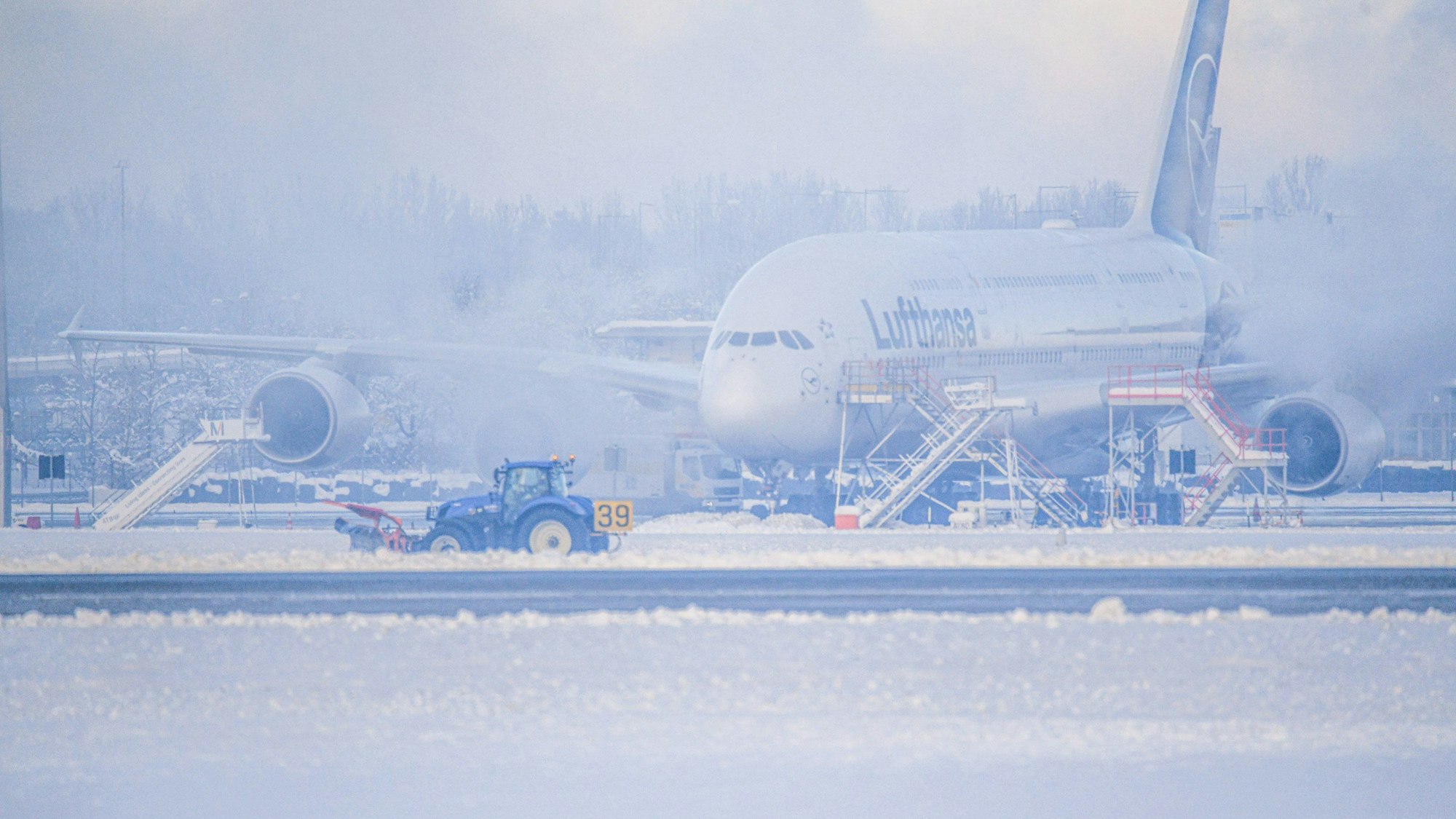 Ein Schneeräumer fährt im Schneetreiben auf dem Flughafen vor einer Maschine entlang. Wegen Eisregen konnten Flugzeuge in München stundenlang nicht starten oder landen.