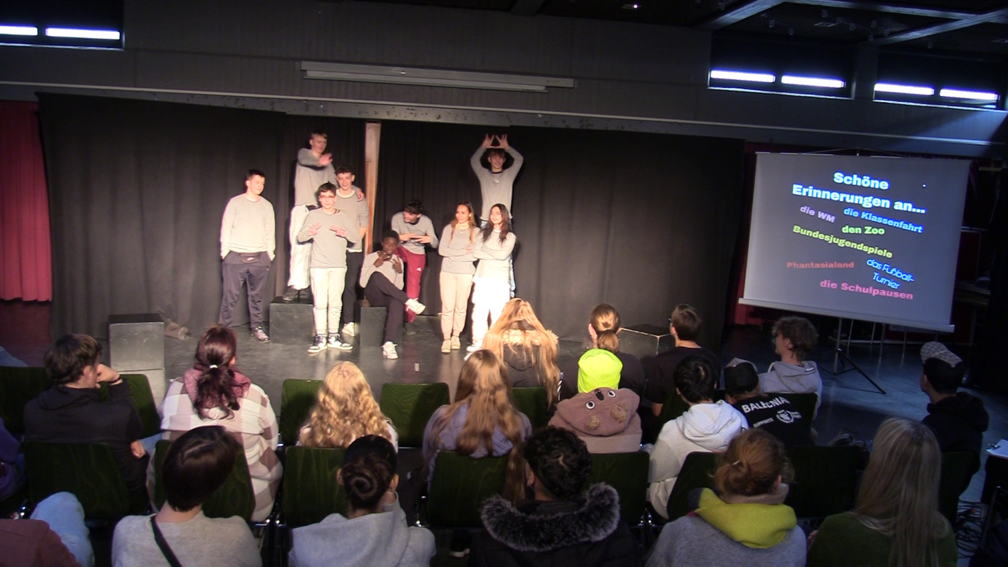 Auf einer Bühne stehen und sitzen mehrere Jugendliche, fotografiert wurde aus dem Publikum heraus. Man sieht Schüler auf Stühlen sitzen.