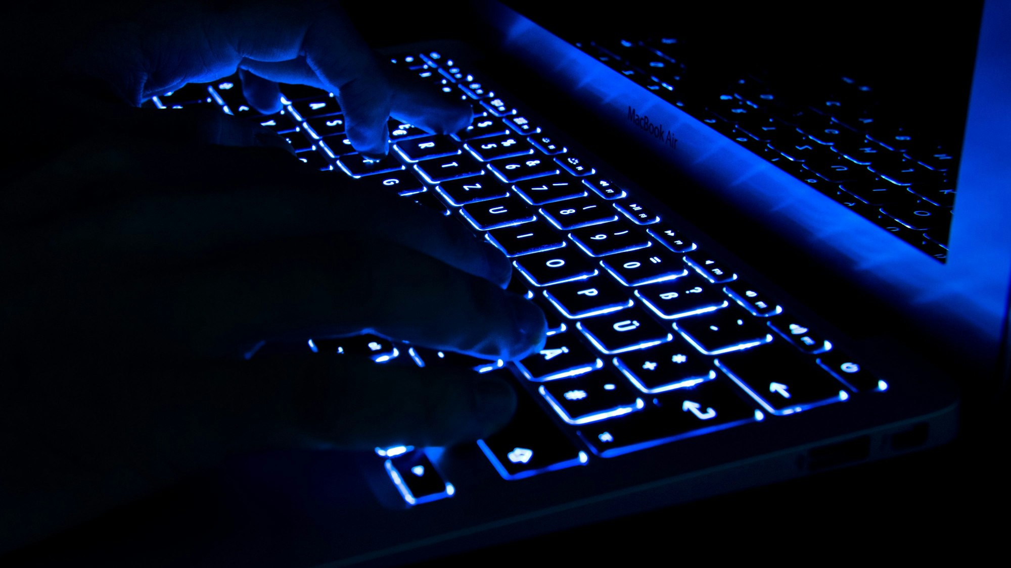 Ein Mann tippt in der Dunkelheit auf erleuchtete Tasten eines Laptops.