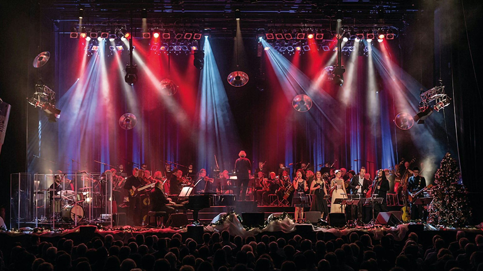 Auf einer rot und blau beleuchteten Bühne stehen Musiker, Sänger und ein Dirigent.