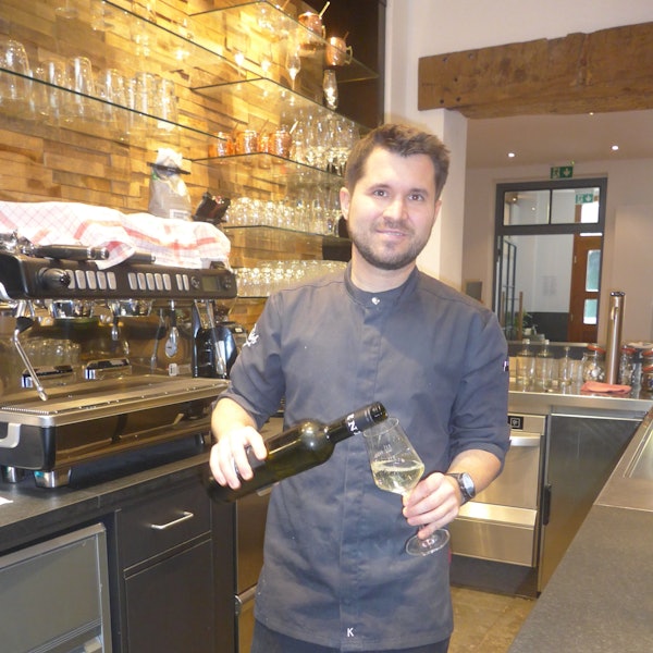 Auf dem Foto ist der Bedburger Gastronom Christof Hölscher zu sehen. Er schenkt Weißwein aus einer Flasche in ein Glas ein.