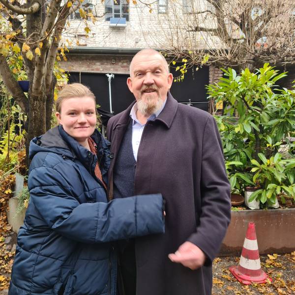 Eine junge Frau und ein älterer Mann mit Bart stehen in einem begrünten Hinterhof.