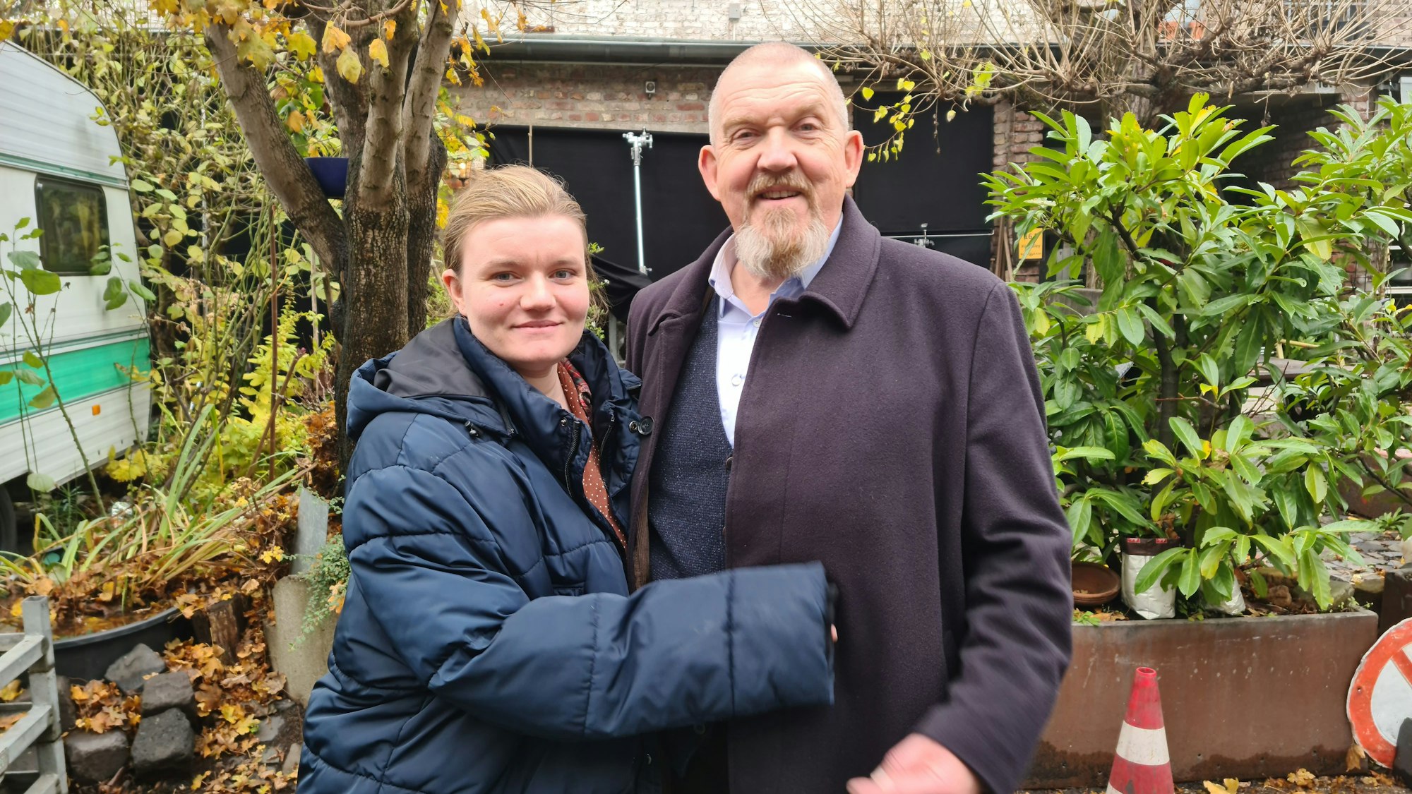 Eine junge Frau und ein älterer Mann mit Bart stehen in einem begrünten Hinterhof.