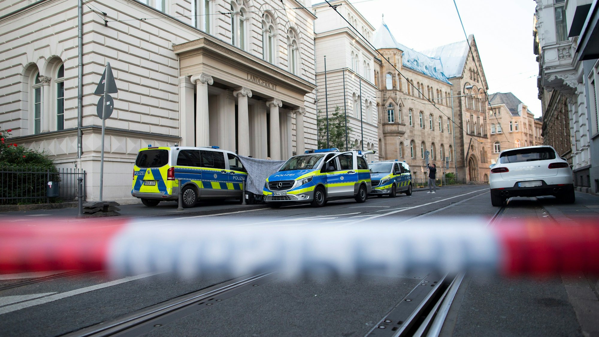 Hinter einem unscharfen Absperrband stehen Polizeiautos vor dem Bonner Landgericht, auf dem Boden sind Schienen der Straßenbahn eingelassen.