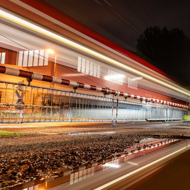 Eine Pfütze spiegelt den Bahnübergang Tannenbergstraße, durch den gerade eine S-Bahn fährt.