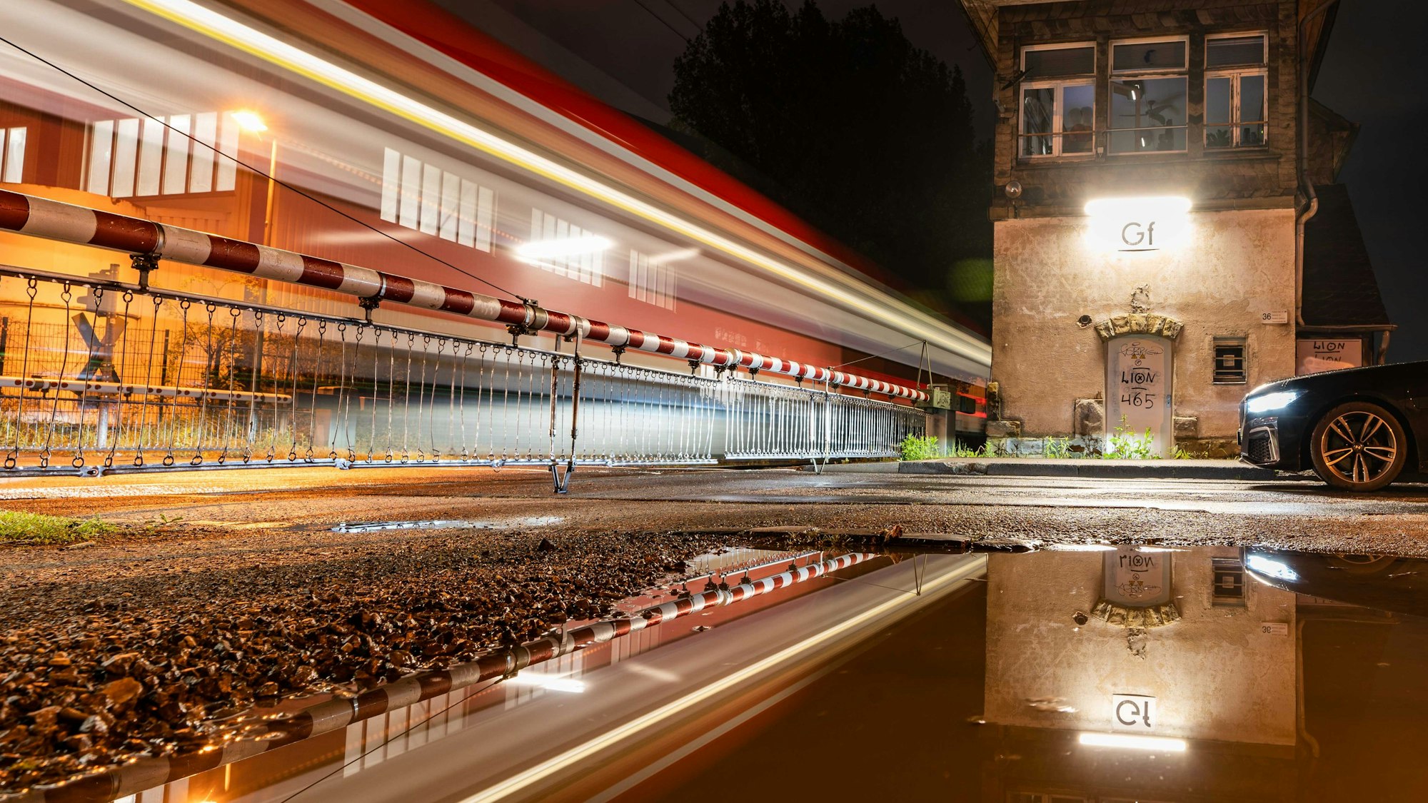 Eine Pfütze spiegelt den Bahnübergang Tannenbergstraße, durch den gerade eine S-Bahn fährt.