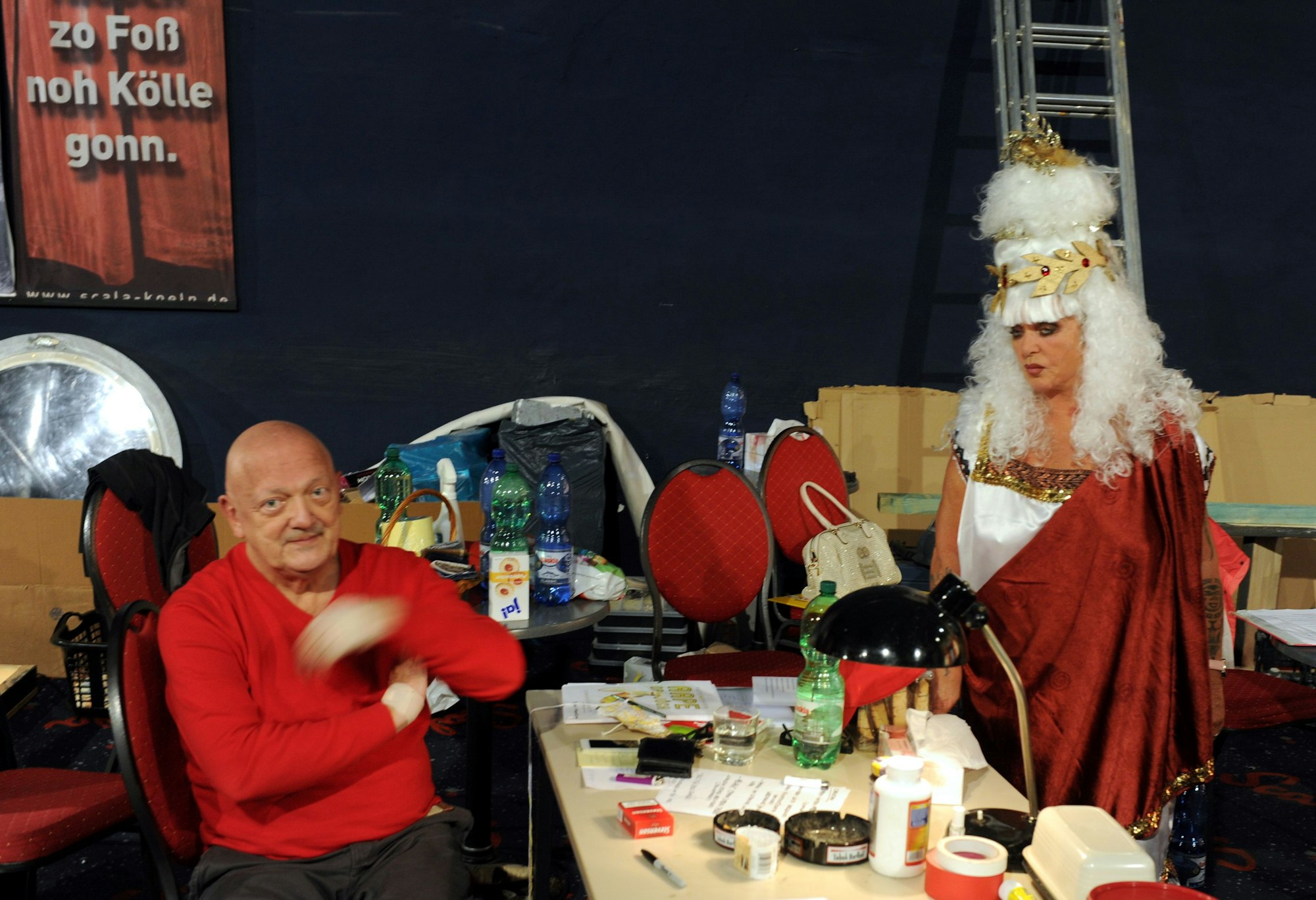 Gigi Herr steht verkleidet vor einem Schreibtisch, an dem Walter Bockmayer sitzt.