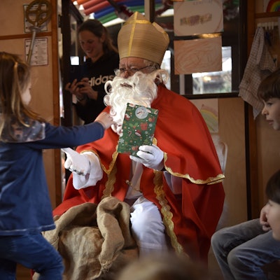 Ein Mann mit rotem Mantel, Mitra auf dem Kopf und langem, weißen Bart hält einem Mädchen eine bunte Geschenk-Tüte hin.