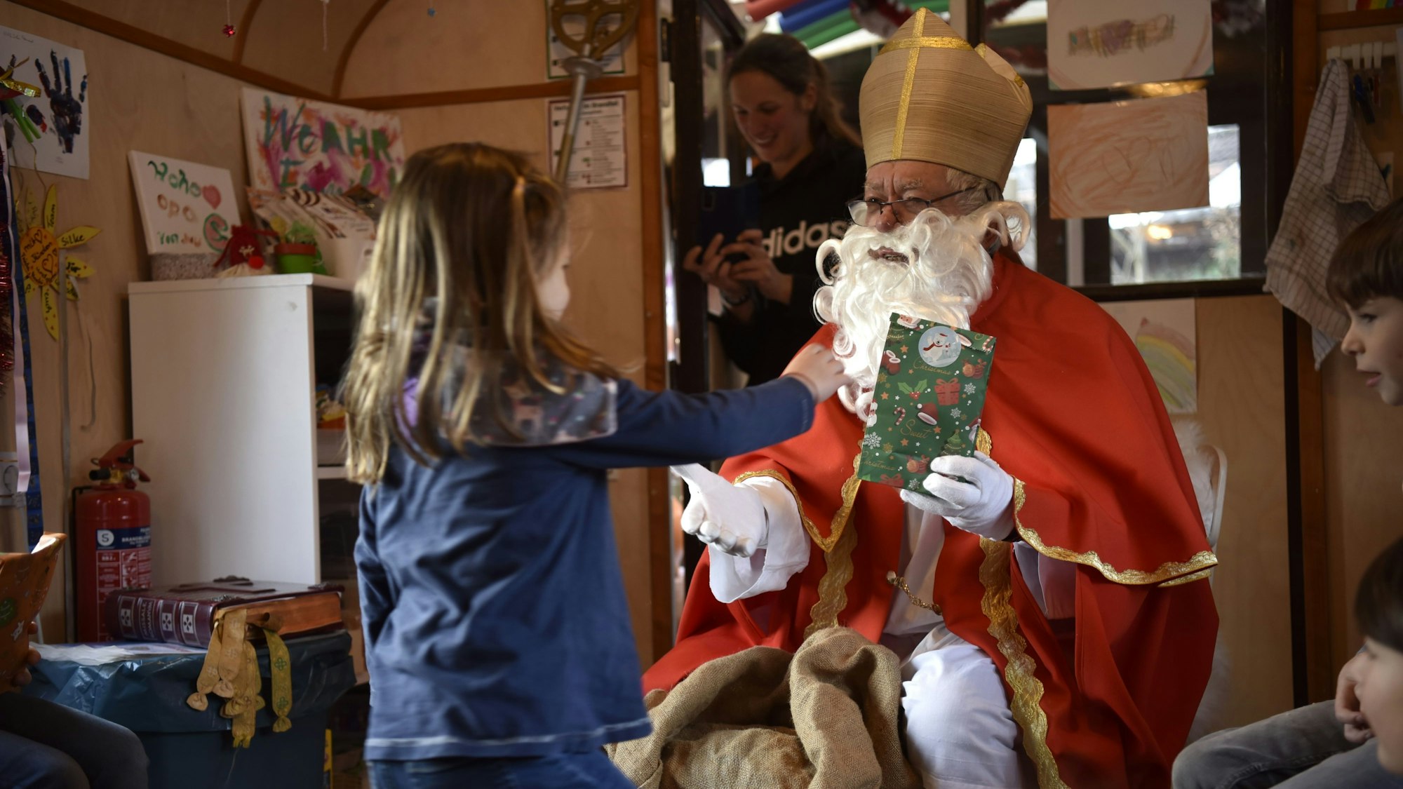 Ein Mann mit rotem Mantel, Mitra auf dem Kopf und langem, weißen Bart hält einem Mädchen eine bunte Geschenk-Tüte hin.