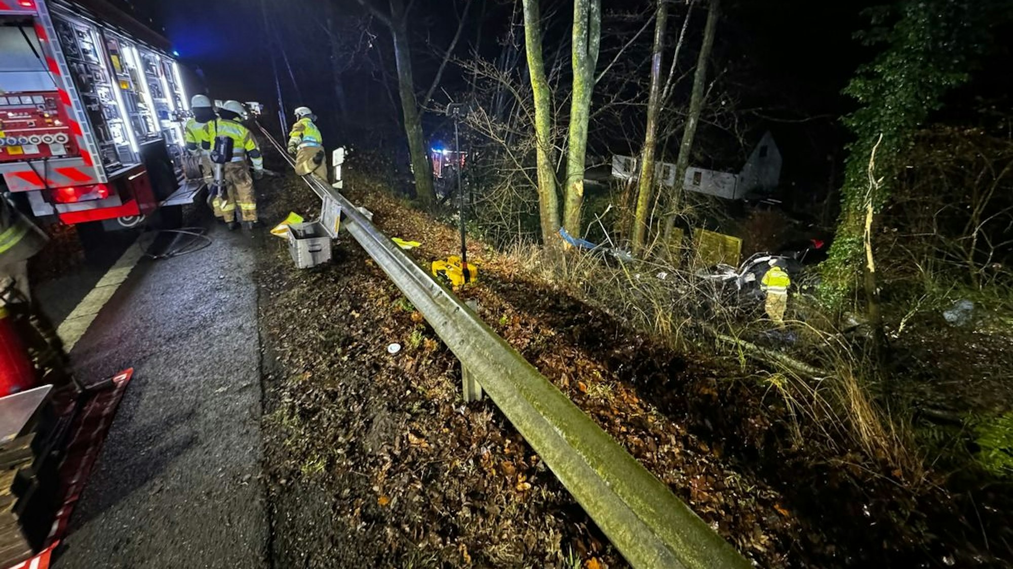 Feuerwehrautos stehen auf der Overather Straße, Einsatzkräfte der Feuerwehr arbeiten an einem Autowrack im Wald dahinter.