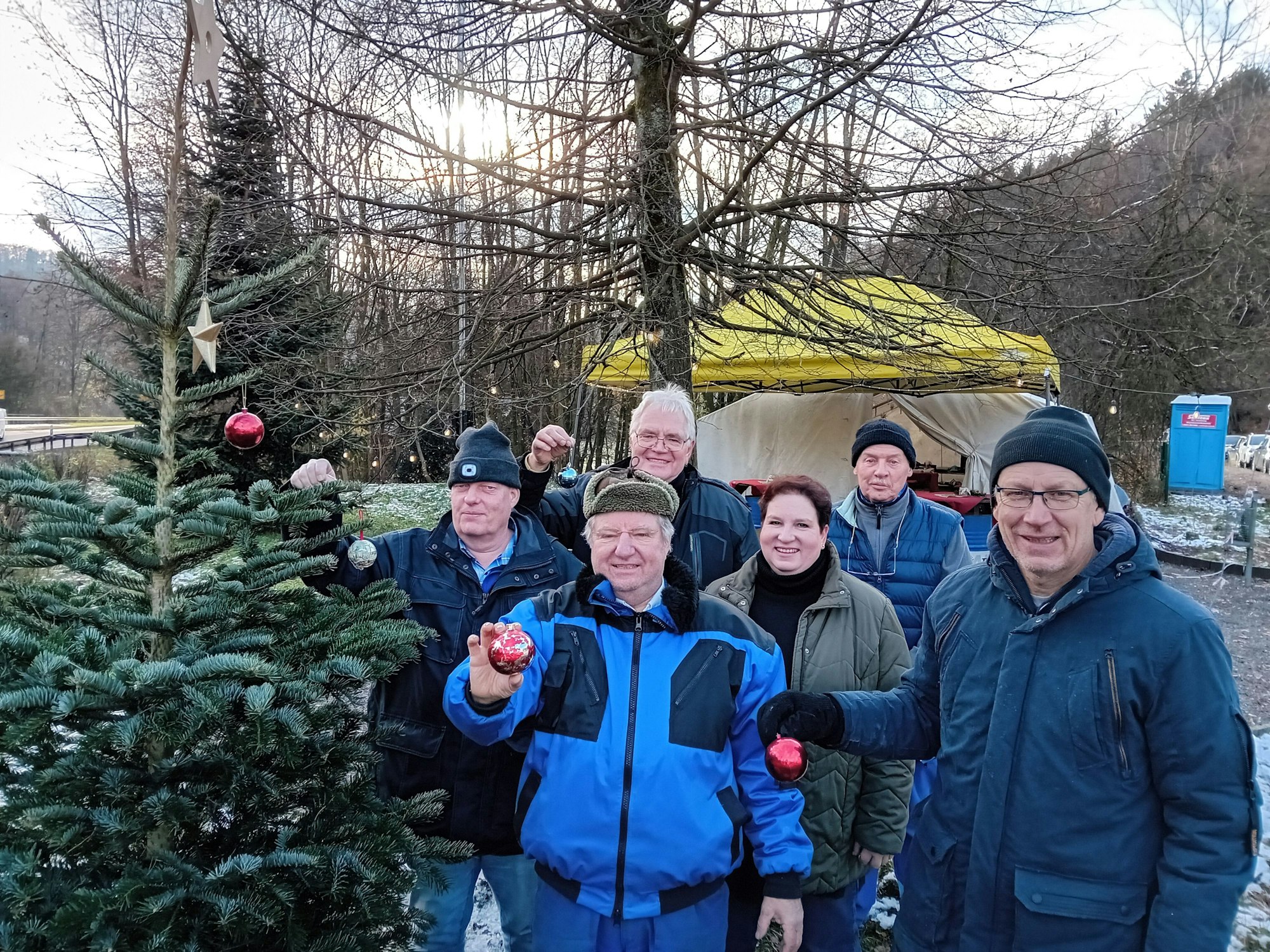 Männer in Arbeitskleidung stehen neben einem Tannenbaum und zeigen Christbaumschmuck.